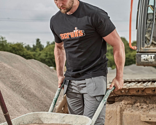 A man wearing a black short-sleeve t-shirt with an orange and white Scruffs logo, working next to a wheelbarrow and an excavator at a construction site.